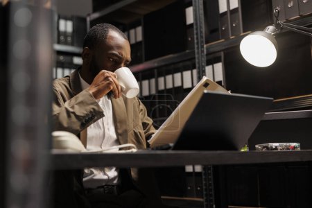 Photo for African american cop drinking coffee and reading csi report at night time. Detective sitting at workplace desk, holding tea mug and studying crime case file in dark office room - Royalty Free Image