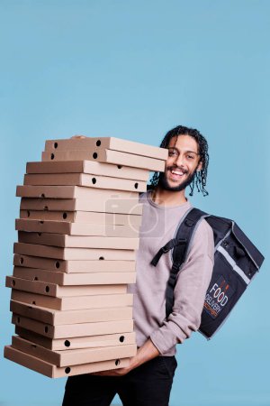 Téléchargez les photos : Livreur arabe insouciant portant de grandes boîtes de pizza pile et regardant la caméra avec expression joyeuse. Service de livraison de restaurant souriant courrier debout énorme pile de paquets de restauration rapide - en image libre de droit