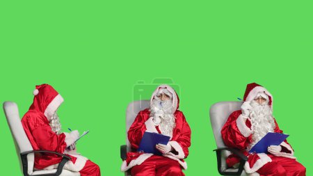 Photo for Santa claus man taking notes on clipboard while he wears seasonal red costume in studio. Person portraying saint nick and writing information on papers, festive winter holiday concept. - Royalty Free Image