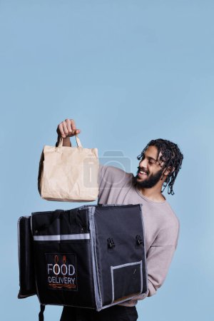 Photo for Happy arab deliveryman holding take out paper bag while delivering restaurant lunch. Cheerful food delivery service courier with carefree expression taking package from thermal backpack - Royalty Free Image