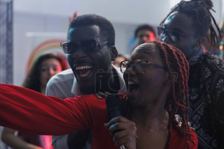 Téléchargez les photos : Femme afro-américaine insouciante chantant au microphone et prenant selfie avec des amis dans une boîte de nuit. Les gens souriants joyeux rient et s'amusent tout en posant pour la photo dans le club - en image libre de droit