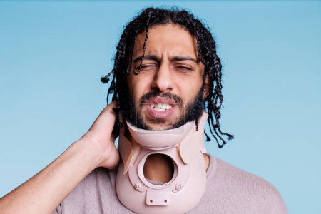 Téléchargez les photos : Homme arabe portant un collier cervical et grimaçant dans la douleur tout en touchant le cou. Jeune blessé avec une expression faciale douloureuse portant une attelle, se rétablissant d'un traumatisme médullaire - en image libre de droit