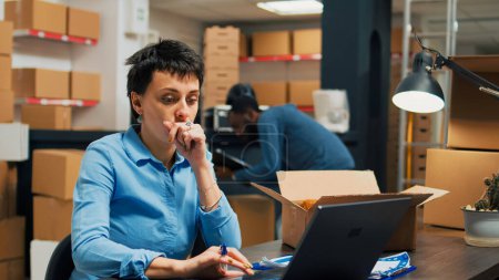 Foto de Woman employee analyzing goods from warehouse shelves, looking at merchandise in cardboard boxes before shipping goods order. Young adult working in storage space with supply chain. - Imagen libre de derechos