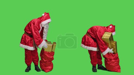 Téléchargez les photos : Père Noël porte un sac avec des cadeaux pour la fête de Noël, la livraison de cadeaux et de jouets aux enfants du monde entier. Homme dépeignant Père Noël et tenant sac rouge avec des paquets. - en image libre de droit