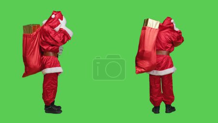 Photo for Saint nick with migraine carries sack, trying to deliver gifts boxes to children on time for christmas eve holiday. Unwell main character having painful headache, full body greenscreen. - Royalty Free Image