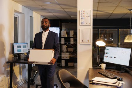 Photo for African american worker packing personal belongings after being fired from corporate job. Depressed executive manager carrying cardboard box while leaving office late at night. Business concept - Royalty Free Image