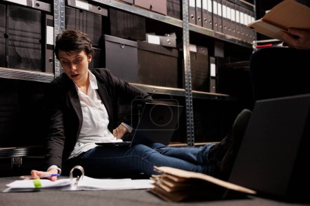 Photo for Overworked inspector sitting on floor in arhive room, analyzing criminology report. Private detectives working overhours at criminal case, checking crime scene evidence trying to catch suspect - Royalty Free Image