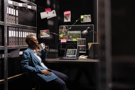 Photo for Pensive african american woman investigator drinking coffee, analyzing murder case. Thoughtful woman holding tea cup, sitting at detective desk while studying clues at night time - Royalty Free Image