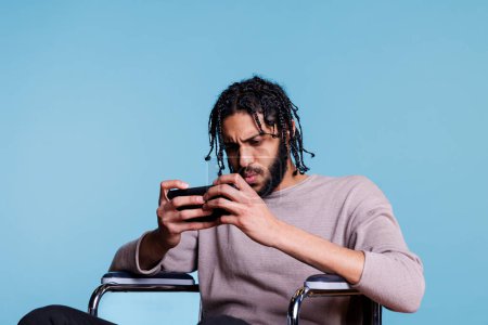 Photo for Focused gamer with disability playing on smartphone with tensed facial expression. Concentrated arab man in wheelchair holding mobile phone and using online game application - Royalty Free Image