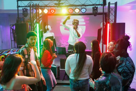 Téléchargez les photos : Clubbers foule dansant au concert de musique techno et faire la fête dans une boîte de nuit. Dj chantant et se produisant en direct sur scène tandis que diverses personnes se déplaçant et s'amusant sur la piste de danse du club - en image libre de droit