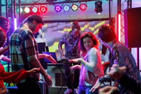Téléchargez les photos : Les gens dansent pendant que le DJ se produit en direct sur scène à la soirée disco de la boîte de nuit. Divers hommes et femmes s'amusent et se déplacent sur la piste de danse lors d'un concert de musicien au club - en image libre de droit