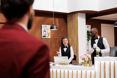 Photo for Receptionist greets hotel guest, providing excellent concierge services for businessman on work trip. Traveller with luggage being welcomed by front desk staff and bellboy, luxury resort. - Royalty Free Image