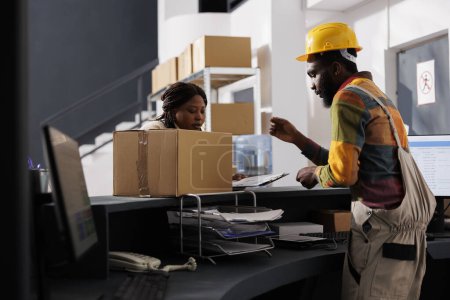 Photo for Courier with clipboard bringing parcel to delivery service and register at postal warehouse reception. African american shipment managers checking package documentation at checkout - Royalty Free Image
