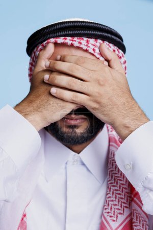 Photo for Man dressed in islamic clothes closing eyes with hands while showing see no evil three wise monkeys sign. Young muslim person showcasing eyesight problem concept closeup in studio - Royalty Free Image
