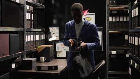Foto de Saliendo de la sala de pruebas, un detective privado cierra su computadora personal. Agente de la ley afroamericano organiza su escritorio, organiza el papeleo, y sostiene su bolso portátil. - Imagen libre de derechos