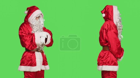 Photo for Adult in costume looks at wristwatch to check time against greenscreen backdrop, wearing santa claus red festive suit. Person acting like saint nick to celebrate winter holiday season. - Royalty Free Image