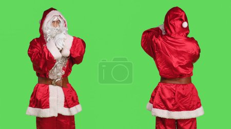 Téléchargez les photos : Impatient Père Noël claus en costume vérifier l'heure sur montre-bracelet alors qu'il se tient sur fond d'écran vert. Jeune adulte habillé en costume de caractère d'hiver festif attendant quelque chose, esprit de Noël. - en image libre de droit