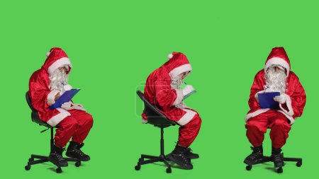 Photo for Winter character taking notes on papers, sitting on chair to write information on clipboard during christmas eve holiday. Santa claus man in cosplay posing over greenscreen backdrop. - Royalty Free Image