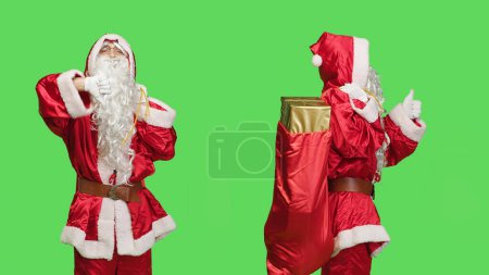 Photo for Santa does thumbs up and thumbs down against greenscreen background, carrying sack of presents on camera. Undecided saint nick shows like and dislike sign, wearing famous costume. - Royalty Free Image