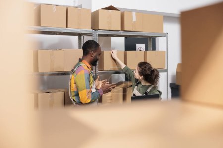 Photo for Multi ethnic storehouse team checking cardboard boxes, working at customers orders before start shipping packages. Employees doing products quality control, discussing inventory report in warehouse - Royalty Free Image