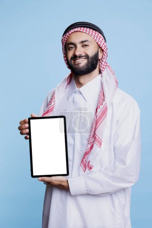 Foto de Hombre musulmán sonriente mostrando tableta digital con pantalla blanca vacía y mirando a la cámara con expresión alegre. Feliz árabe anunciante sosteniendo retrato de pantalla táctil en blanco - Imagen libre de derechos