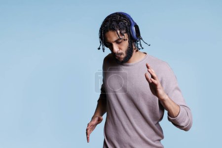 Téléchargez les photos : Homme arabe concentré dansant tout en écoutant de la musique dans les écouteurs. Jeune axé sur les chansons playlist dans les écouteurs sans fil déplacer les mains et le corps sur fond bleu - en image libre de droit