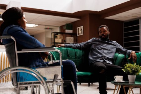 Junger afroamerikanischer Mann spricht mit Freundin im Rollstuhl, während er zusammen in Hotellobby sitzt und mit behinderter Frau ins Ausland reist. Menschen mit Behinderungen und Reisen