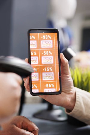 Close up of male hand holding smartphone with Black Friday discount coupon while paying for purchases at checkout. Cashier scanning promotional promo code displayed on consumer mobile device