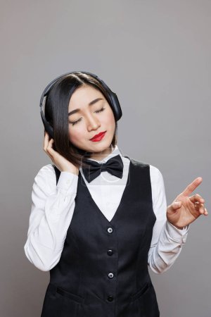 Téléchargez les photos : Femme asiatique réceptionniste en uniforme jouissant playlist dans les écouteurs. Jeune serveuse attrayante avec les yeux fermés écoutant de la musique dans des écouteurs sans fil tout en posant en studio - en image libre de droit