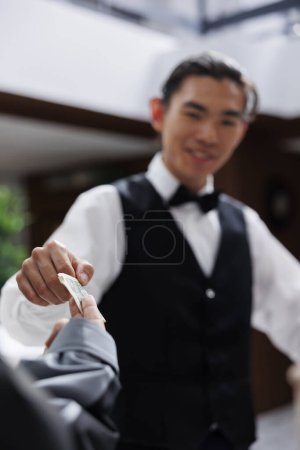 Photo for Photo showcasing happy asian bellboy receiving cash tip from tourist after assisting with luggage in hotel lobby. Guest checks in receives friendly service and gives money tip to concierge. - Royalty Free Image