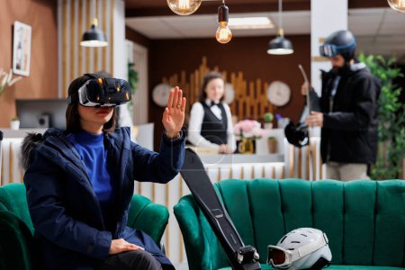 Photo for Asian lady wearing digital VR headset having skiing equipment and winter jacket in hotel lobby. Female tourist searching with vr glasses for best slopes at ski mountain resort snow adventure. - Royalty Free Image