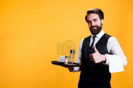 Photo for Experienced butler gives thumbs up in studio, presenting agreement symbol and being positive on camera. Young man with wiater occupation shows like approval before serving cup of coffee. - Royalty Free Image