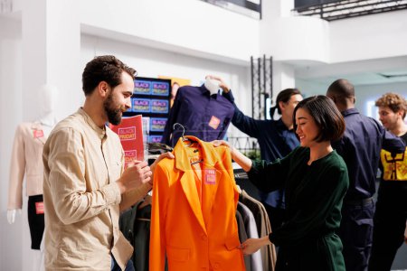 Photo for Happy diverse couple shopping on Black Friday in clothing store, looking at orange formal jacket with red sale tag and discussing price. Husband helping wife to choose clothes during seasonal sales - Royalty Free Image