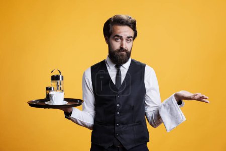 Photo for Male waiter feeling unsure in studio, doing i dont know gesture and shurgging. Restaurant employee acting clueless and unsure while he is holding platter in hand, poses over yellow background. - Royalty Free Image