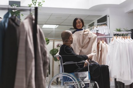 Photo for Clothing store smiling asian woman consultant helping arab man with disability in exploring formal womenswear rack. Seller assisting buyer in wheelchair in selecting beige jacket - Royalty Free Image