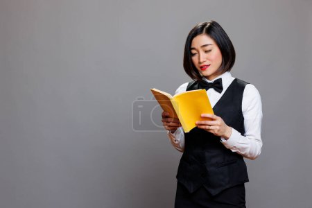 Téléchargez les photos : Serveuse asiatique réfléchie vêtue d'un livre de lecture uniforme professionnel avec couverture jaune. Jeune femme séduisante réceptionniste tenant un journal à couverture souple tout en posant en studio - en image libre de droit