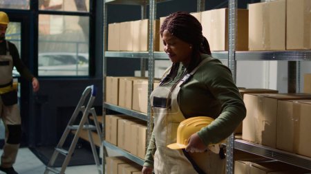 Foto de Retrato del alegre trabajador de almacén afroamericano parado frente a estantes llenos de mercancías empaquetadas en cajas de cartón listas para ser enviadas, feliz de trabajar en un entorno profesional - Imagen libre de derechos
