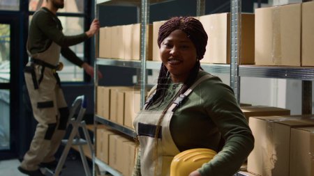 Téléchargez les photos : Portrait de joyeux ouvrier d'entrepôt afro-américain debout devant des étagères remplies de marchandises emballées dans des boîtes en carton prêtes à être expédiées, heureux de travailler dans un environnement professionnel - en image libre de droit