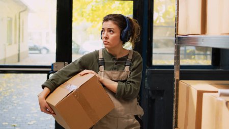 Photo for Employee moving stock boxes on racks, listening music on headset while she prepares retail orders for delivery. Young woman working on products shipment, showing dance moves in depot. - Royalty Free Image