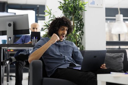 Téléchargez les photos : Entrepreneur arabe excité réjouissant campagne de marketing réussie dans le bureau de démarrage d'entreprise. Employé d'entreprise montrant geste gagnant tout en vérifiant le rapport de vente sur ordinateur portable - en image libre de droit