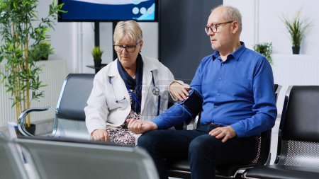 Téléchargez les photos : Médecin principal mesurant l'hypertension du patient à l'aide d'un tonomètre, vérifiant l'hypotension lors d'un rendez-vous dans la salle d'attente de l'hôpital. Homme âgé ayant un examen cardiaque - en image libre de droit