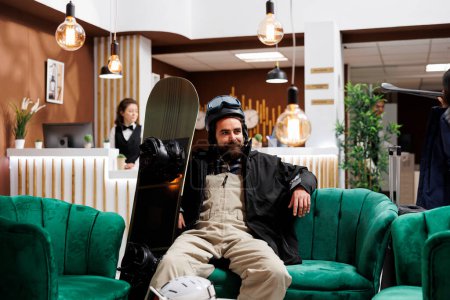 Téléchargez les photos : Jeune homme caucasien en tenue de sport d'hiver assis confortablement dans le salon de l'hôtel exclusif en attente d'enregistrement. Touriste masculin actif à l'accueil de la station de vacances en attente d'hébergement. - en image libre de droit