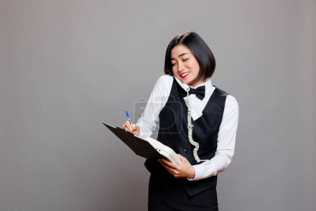 Photo for Smiling asian waitress listening to manager instructions on landline phone and taking notes in clipboard. Catering service woman worker speaking on telephone and writing checklist - Royalty Free Image