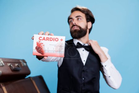 Photo for Hotel concierge presents cardiac pills in front of studio camera, recommending medication for heart disease or problems. Young man doorkeeper shows box of medicaments treatment. - Royalty Free Image
