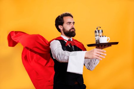 Photo for Professional restaurant waiter character holds dish tray and towel while he wears red cape fluttering in studio. Young adult posing with strength and confidence, serving food on platter. - Royalty Free Image