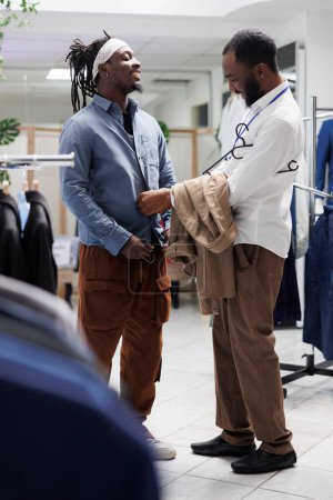 Téléchargez les photos : Souriant homme afro-américain choisissant des vêtements pour hommes avec employé de magasin de vêtements. Chemise de fermeture assistante de centre commercial sur le client pour vérifier la taille et le style dans la boutique de mode - en image libre de droit