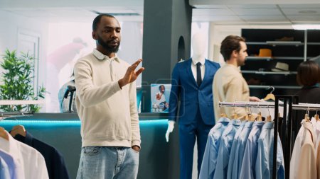 Téléchargez les photos : Homme afro-américain regardant l'hologramme en magasin, en utilisant l'image holographique de réalité augmentée dans la boutique de vêtements de mode. Client masculin vérifiant les vêtements dans le centre commercial. Trépied. - en image libre de droit