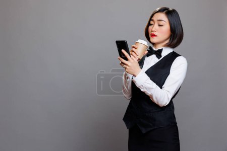 Photo for Asian waitress browsing social media using smartphone during coffee break. Serious woman receptionist wearing uniform texting on mobile phone and holding drink to go paper cup - Royalty Free Image