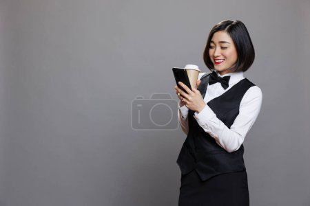Téléchargez les photos : Femme asiatique souriante portant uniforme réceptionniste textos et défilement des médias sociaux tout en buvant du café à emporter. Restaurant serveuse joyeuse profiter du thé dans une tasse en papier et en utilisant un téléphone mobile - en image libre de droit