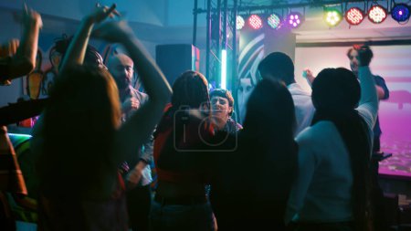 Foto de Jóvenes adultos bailando en la música electrónica, divirtiéndose junto con DJ en el escenario. Hombres y mujeres alegres saltando en la pista de baile bajo luces de discoteca, disfrutando de una fiesta de baile. Disparo de mano. - Imagen libre de derechos